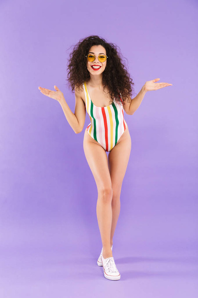 Full length photo of trendy attractive woman with curly hair in swimsuit and sunglasses laughing at camera isolated over violet background in studio - Valokuva, kuva
