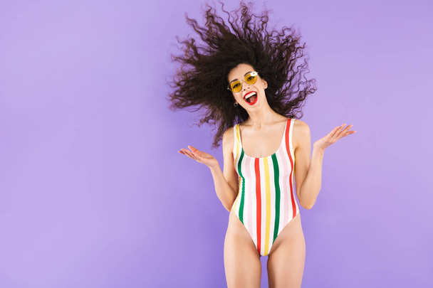 Portrait of astonished caucasian woman in swimsuit and sunglasses fooling around with shaking hair isolated over violet background in studio - Zdjęcie, obraz
