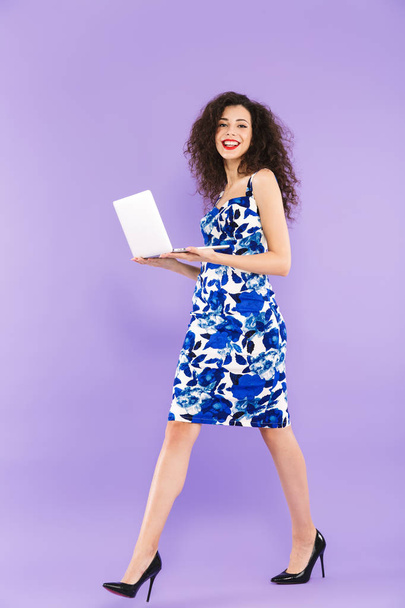 Full length photo of beautiful adult woman with curly hair in dress smiling and walking with laptop in hands isolated over violet background in studio - Foto, afbeelding