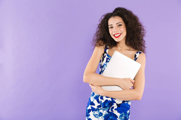Image of beautiful adult woman with curly hair in dress smiling and holding laptop in hands isolated over violet background in studio - Foto, imagen