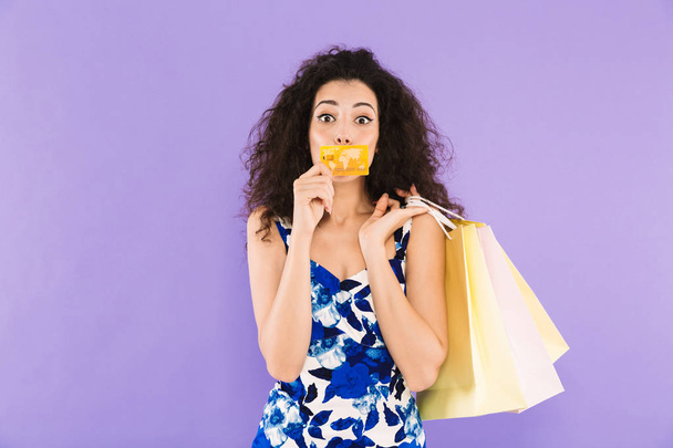 Portrait of shopaholic woman 20s wearing dress holding credit card and paper shopping packages standing isolated over violet background - Foto, immagini