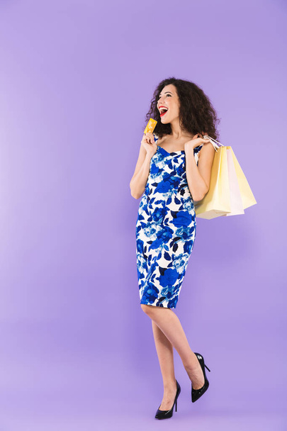 Full length portrait of lucky charming woman 20s wearing dress holding credit card and paper shopping bags standing isolated over violet background - Фото, зображення