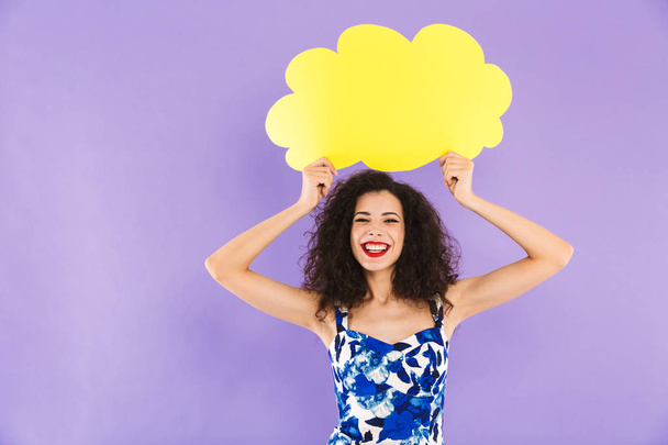 Image of charming happy woman with curly hair in dress smiling and holding blank bubble above her head with copyspace for your text isolated over violet background in studio - Zdjęcie, obraz
