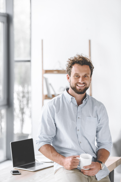 glimlachend zakenman met kop koffie zittend op tafel met laptop en smartphone in kantoor - Foto, afbeelding