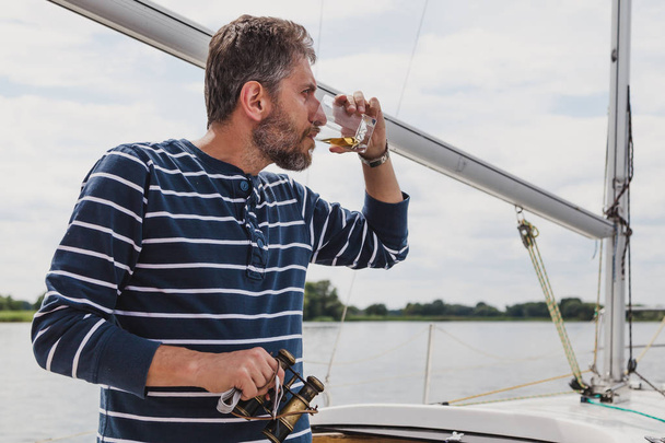 Sea Wolf. Ο καπετάνιος με ένα ριγέ πουλόβερ με μια γενειάδα πίνει ουίσκι σε ένα γιοτ. Στο δεξί του χέρι κρατά ένα παλιό ζευγάρι κιάλια - Φωτογραφία, εικόνα