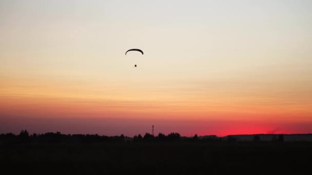 Ο πιλότος σε ένα αλεξίπτωτο που πετά από την κάμερα που μετακινούνται βαθμιαία μακριά στην απόσταση κατά το ηλιοβασίλεμα όμορφο ουρανό. Πανέμορφο φόντο εικόνα φόντου. έννοια της ελευθερίας - Πλάνα, βίντεο