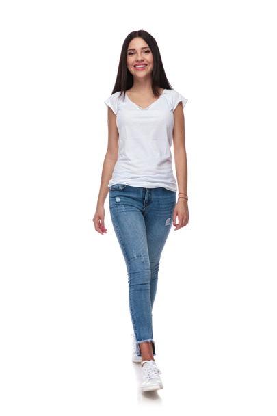 joyful casual woman wearing jeans walking on white background while smiling, full body picture - Photo, Image