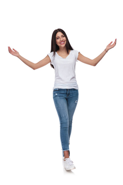 smiling brunette woman stepping on white background and making a welcoming gesture with both hands, full length picture - Photo, Image