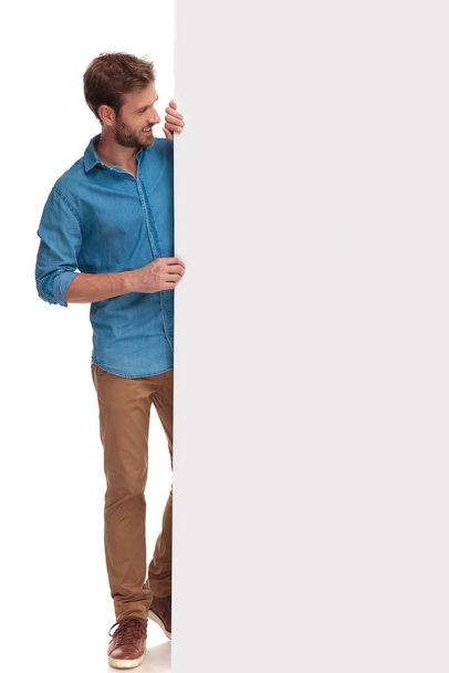 smiling casual man standing behind big blank billboard and showing it on white background - Foto, Bild