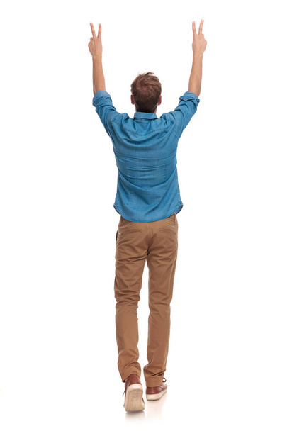 Rückansicht eines lässigen Mannes, der mit den Händen in der Luft geht und das Siegeszeichen auf weißem Hintergrund macht - Foto, Bild