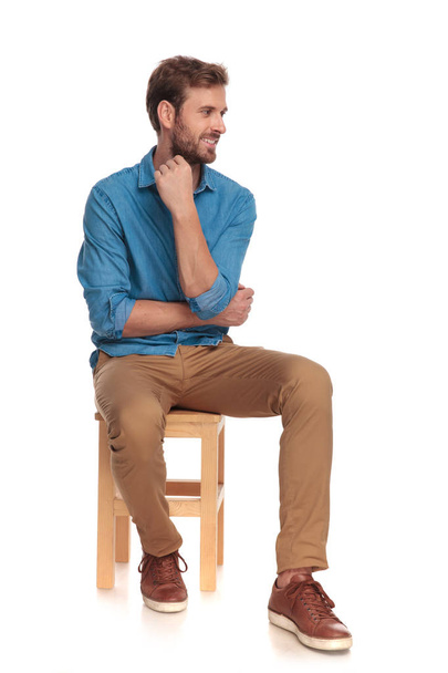 pensive young casual man looks to side while sitting on wooden chair on white background - Zdjęcie, obraz