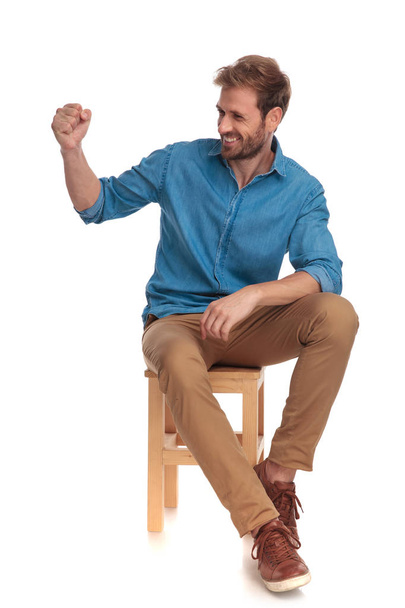 Zijaanzicht van een zittende casual man lachen met vuist omhoog in de lucht op witte achtergrond - Foto, afbeelding