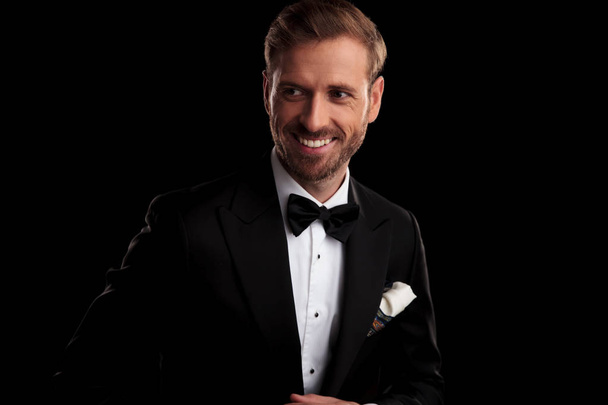 happy elegant man in tuxedo and bowtie looks to side away from the camera on black studio background - Фото, зображення