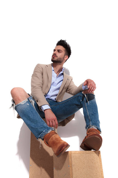 attraktiver smarter, lässiger Mann, der auf Holzkisten sitzt und zur Seite schaut, während er zerrissene Jeans und braune Anzugjacke trägt - Foto, Bild