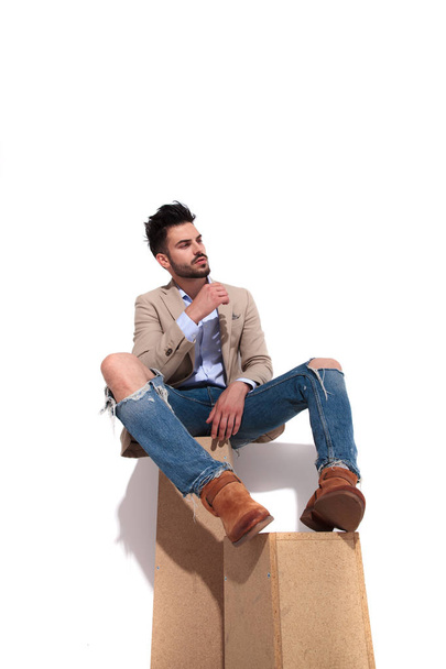 relajado hombre casual inteligente en traje marrón mirando a un lado mientras está sentado en cajas de madera sobre fondo blanco
 - Foto, Imagen