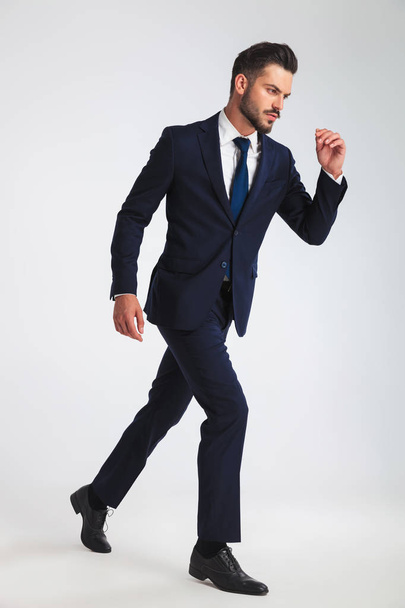 side view of confident businessman in navy suit stepping on light grey background, full length picture - Zdjęcie, obraz