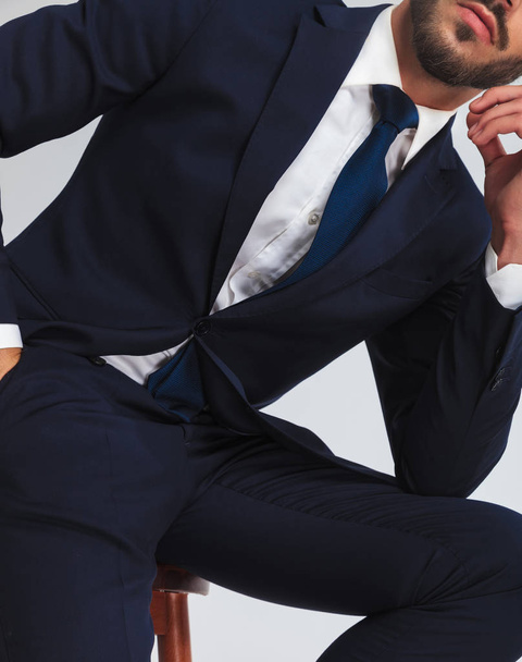 σώμα χαλαρό επιχειρηματία στο Ναυτικό κοστούμι κάθεται σε καρέκλα με το χέρι στην τσέπη στο ανοιχτό γκρι φόντο και κλίνει προς την πλευρά - Φωτογραφία, εικόνα