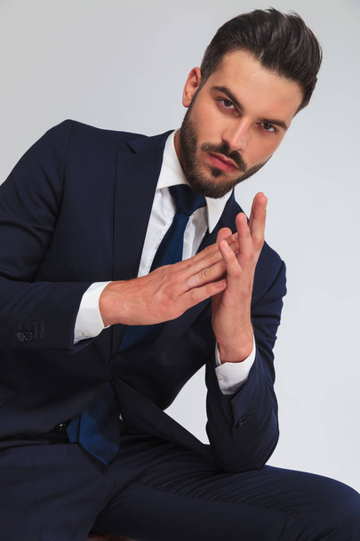 atractivo hombre de negocios en traje azul marino que mantiene las palmas juntas mientras está sentado sobre un fondo gris claro, retrato de imagen
 - Foto, Imagen