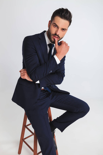 portrait of sexy businessman sitting on a wooden stool and touching his lips on light grey background - Fotografie, Obrázek