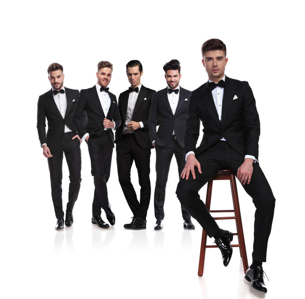 group of five elegant young men in black tuxedoes with leader sitting on chair in front of them, on white background, full body picture - Photo, Image