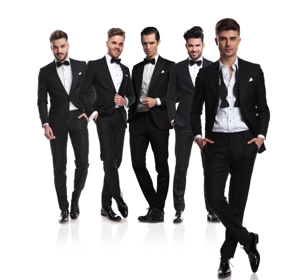 five groomsmen in black tuxedoes with leader with undone collar standing in front cross-legged, on white background, full body picture - Photo, Image