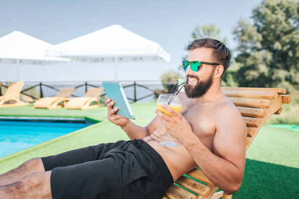 Relaxed and happy guy sits on sunbed and smiles. He holds tablet and looks on it. Man wears sunglasses. He holds cocktail in left hand. He is sitting besides swimming pool. Guy is satisfied. - Photo, image