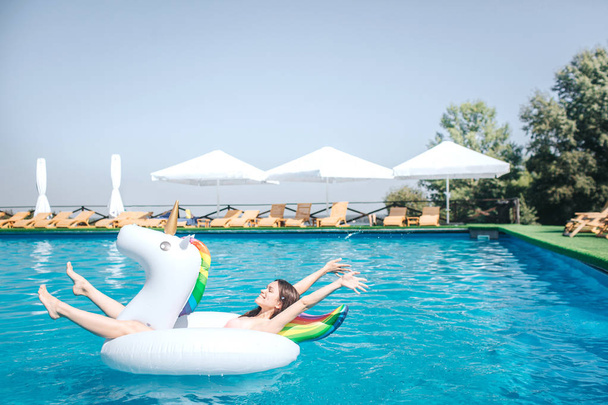 Happy and satisfied girl is lying on air mattress in the middle of swimming pool. She keeps hands and legs in air. Girl is relaxing. She has some rest. - Fotoğraf, Görsel