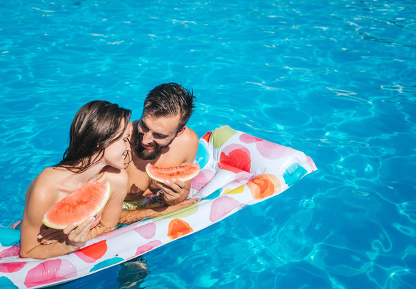 Cute picutre of young man and woman leaning to air mattress and hold pieces of watermelon. They look at eac other and smile. Couple is in swimming pool. - 写真・画像