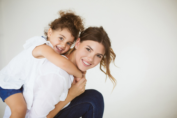 Beautiful smiling mother holding little cute daughter on back happily looking in camera together over white background - Foto, imagen