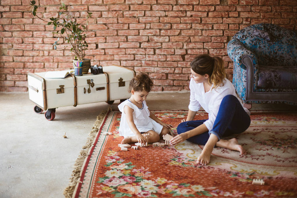 Jonge moeder in wit overhemd en een schattig dochtertje in witte jurk dromerig spelen met houten speelgoed samen op tapijt gezellig thuis - Foto, afbeelding