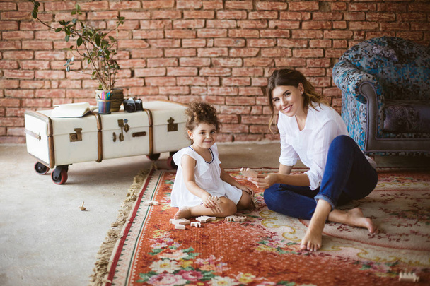 Jovem mãe bonita em camisa branca e pequena filha bonita em vestido branco olhando alegremente na câmera brincando com brinquedos de madeira juntos no tapete em casa aconchegante
 - Foto, Imagem