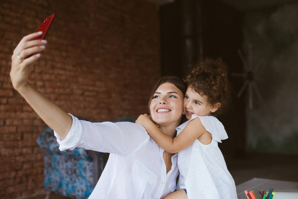 Jonge moeder en schattig dochtertje knuffelen gelukkig nemen van foto's op mobiel op gezellig thuis - Foto, afbeelding