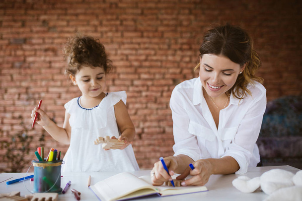 Hermosa madre joven en camisa blanca y su hija bebé en vestido felizmente dibujo en bloc de notas con plumas de fieltro juntos en casa acogedora
 - Foto, Imagen