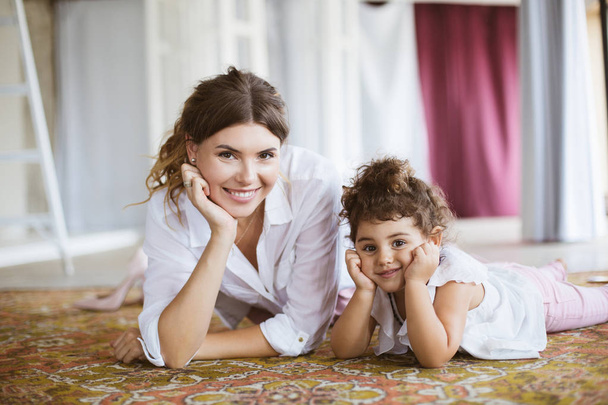 Joven madre sonriente y bonita hija pequeña en camisas blancas mirando felizmente en la cámara acostada en la alfombra juntos en casa
 - Foto, imagen