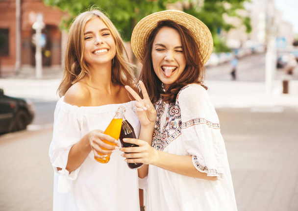 Two young stylish smiling hippie brunette and blond women models in summer sunny day in white hipster clothes posing on the street background. Drinking soda from bottle - Foto, immagini