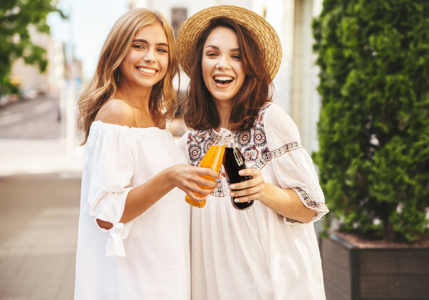 zwei junge, stylisch lächelnde Hippie-Brünette und blonde Models an einem sonnigen Sommertag in weißen Hipster-Klamotten, die auf der Straße posieren. Limo aus der Flasche trinken - Foto, Bild