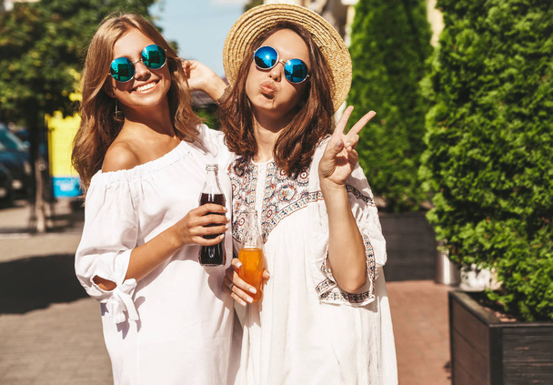 Two young stylish smiling hippie brunette and blond women models in in white summer hipster clothes posing on the street background. Drinking soda from bottle and showing tongue and peace sign - Φωτογραφία, εικόνα