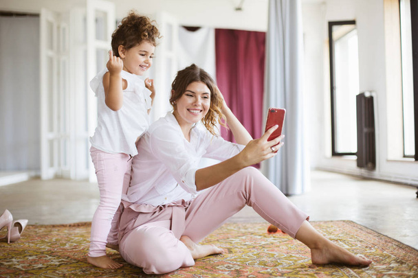 Neşeli anne ve sevimli küçük kız mutlu evde halı üzerinde birlikte vakit cep telefonu üzerinde fotoğraf çekmek - Fotoğraf, Görsel