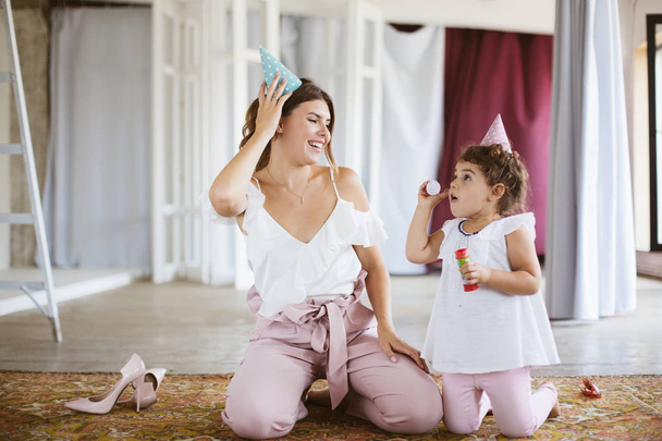 Молодая улыбающаяся мать и милая маленькая дочка в шапочках на день рождения с радостью пускают пузырьки проводить время вместе на ковре дома
 - Фото, изображение