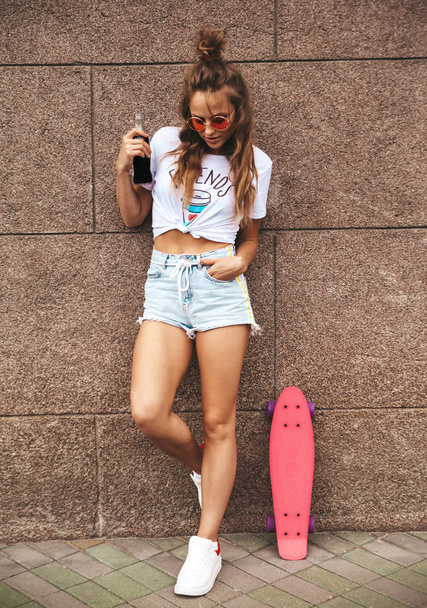 Beautiful cute blond teenager model without makeup in summer hipster white clothes with pink penny skateboard posing near wall on the street.Drinking colorful cocktail from bottle - Фото, изображение