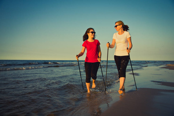 Pretty women walking together with tourist sticks at seaside. Nordic walking. - Photo, image