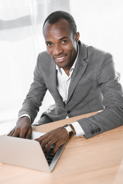 улыбающийся африканский мужчина смотрит в камеру, используя ноутбук на столе
  - Фото, изображение