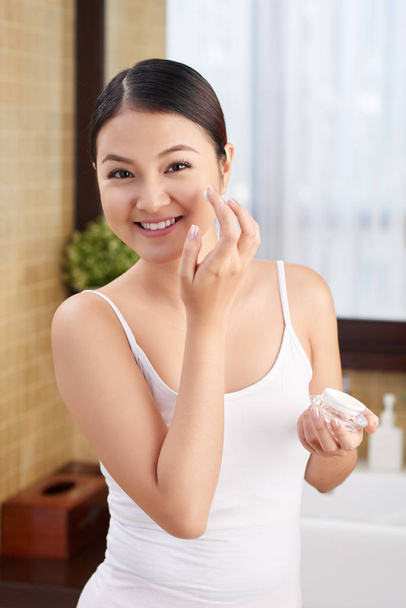 Cheerful young Asian woman applying hydration lotion on her face - Foto, Imagen