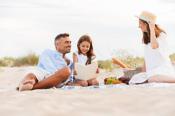 Happy family with father, mother, daughter having picnic at the beach, reading book - Фото, изображение