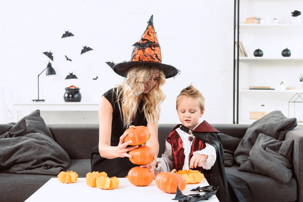 portrait of mother and son in halloween costumes sitting at coffee table with pumpkins at home - Photo, Image