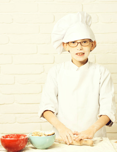 young boy cute cook chef in white uniform and hat on stained face flour with glasses cooking on table with rolling pin and colorful bowls with chocolate cookies on brick wall background - Foto, Imagen