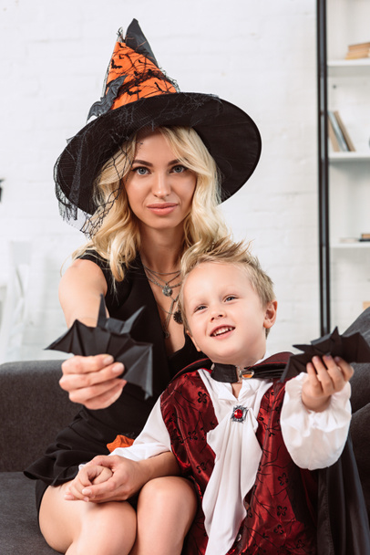 mother and little son in halloween costumes showing black paper bats on sofa at home - Valokuva, kuva