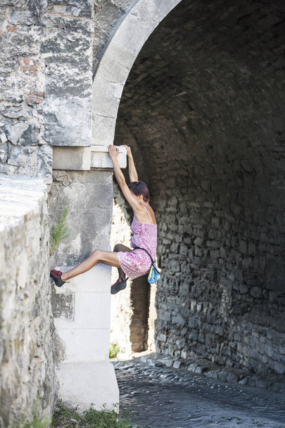 Kobieta w sukience jest wspinaczka mur z cegły. Dziewczyna wspina się na ogrodzenie z kamienia. Pnącze na danej ulicy miasta. Na ścianie starego zniszczonego budynku. - Zdjęcie, obraz