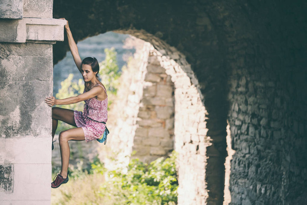 The girl climbs the stone wall. A woman in a summer dress climbs the wall of an old destroyed building. Brick fence. The climber is hanging on the city building. Strengthening the ruined castle. - Photo, Image