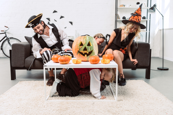 jongetje verbergen van ouders en zus in halloween kostuums onder tafel met pompoenen thuis - Foto, afbeelding